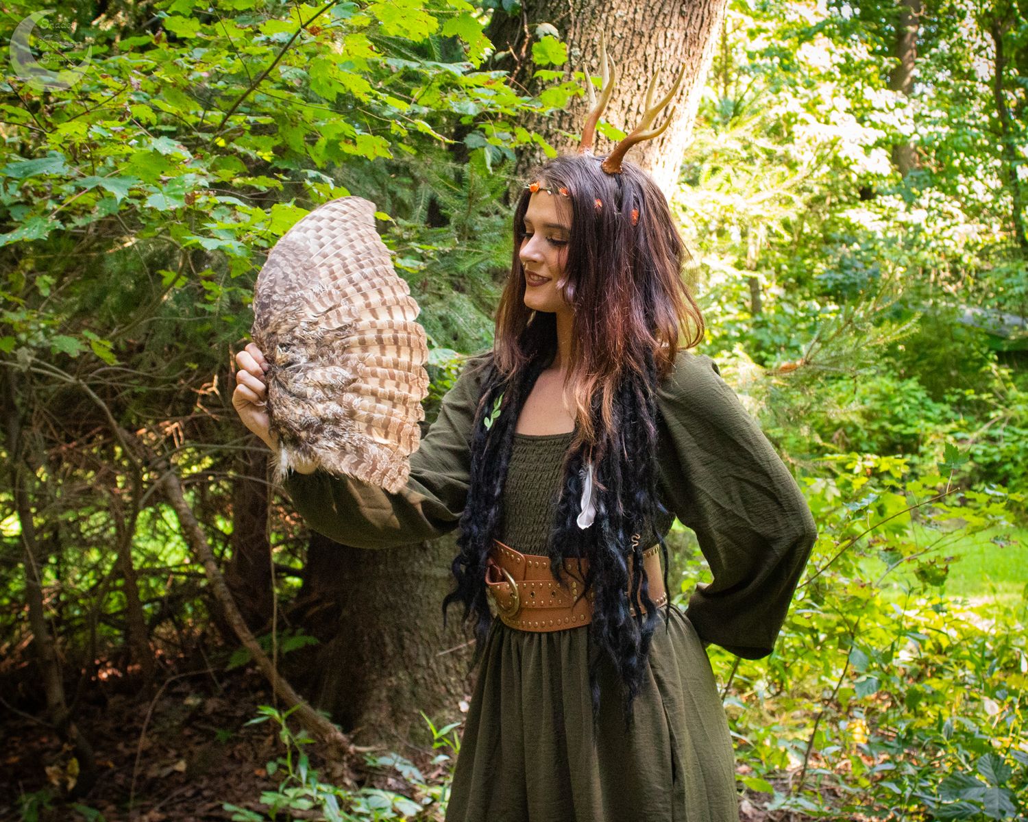 Figure in dark dress poses elegantly against lush forest backdrop with dappled sunlight.