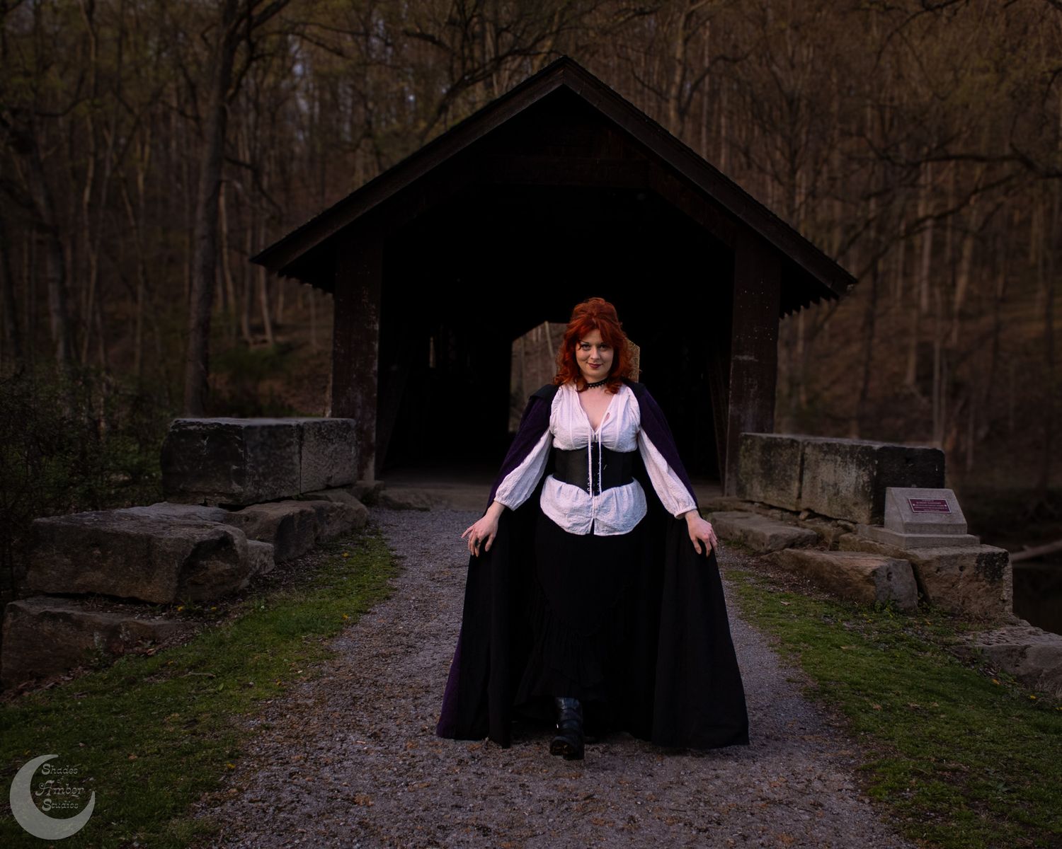 A figure in gothic attire stands before a dark wooden pavilion on a misty forest path.