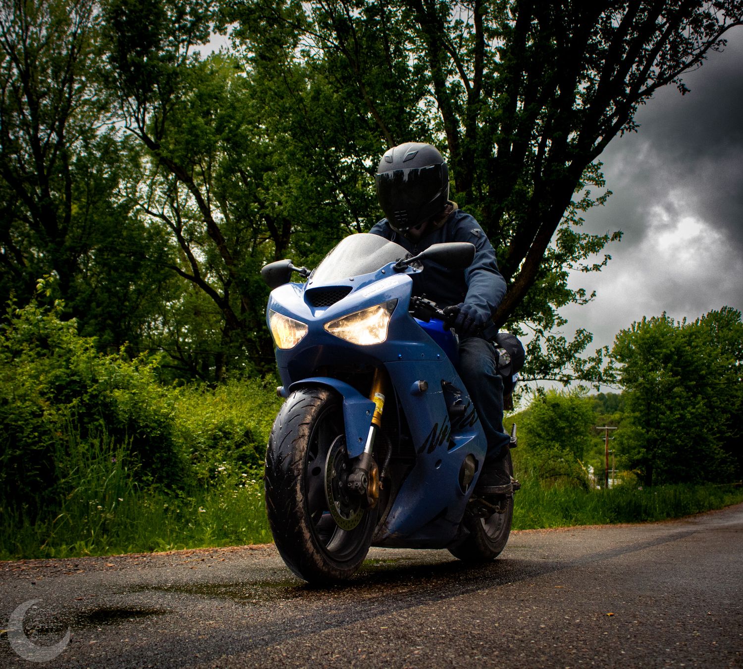 A blue sportbike motorcycle parked on a dirt road under dramatic storm clouds with trees in the background.
