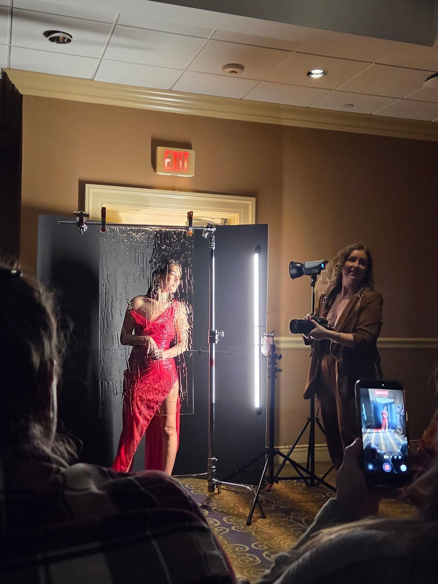 Behind-the-scenes photo shoot in a hotel hallway shows photographer capturing model in red dress against dramatic lighting setup.