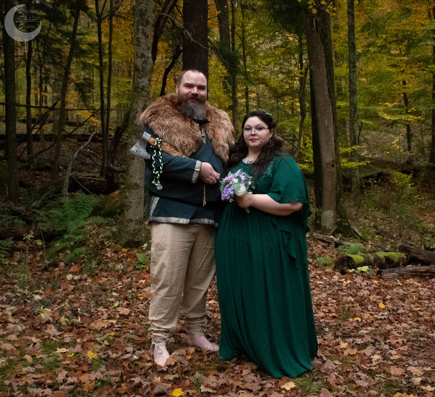 A couple embraces while standing on fallen autumn leaves in a wooded forest setting.