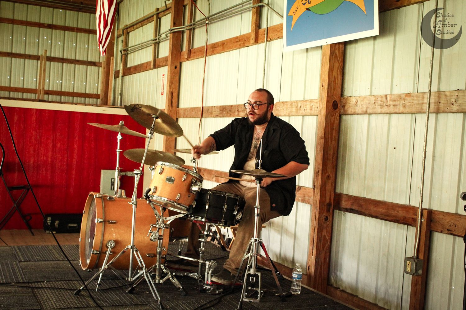 A drummer performs on a vintage drum kit inside a rustic wooden barn-like structure.
