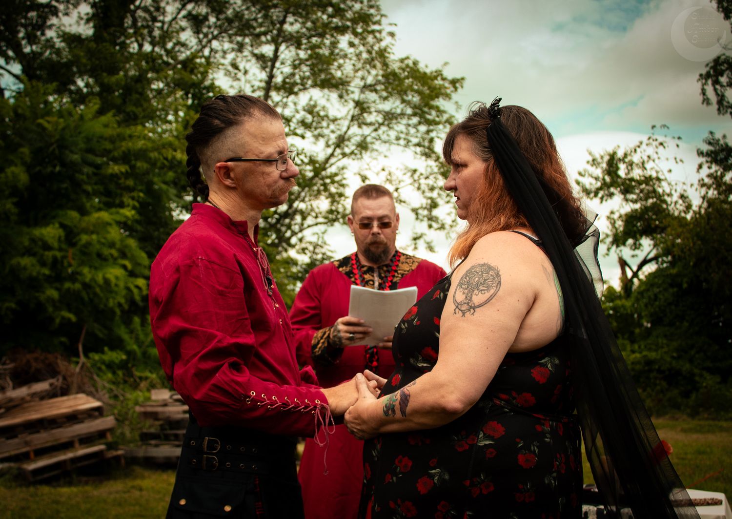 A couple in red and black attire hold hands during an outdoor wedding ceremony in a garden setting.