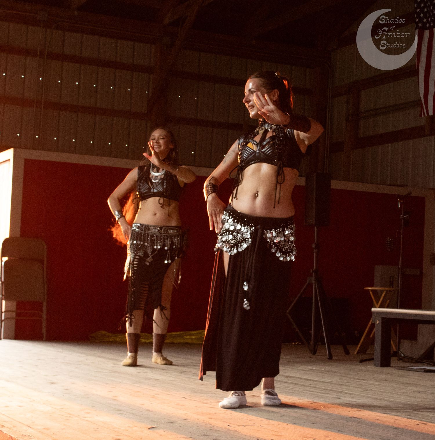 Two dancers in black and silver belly dance costumes perform on stage with decorative coin belts and crop tops.