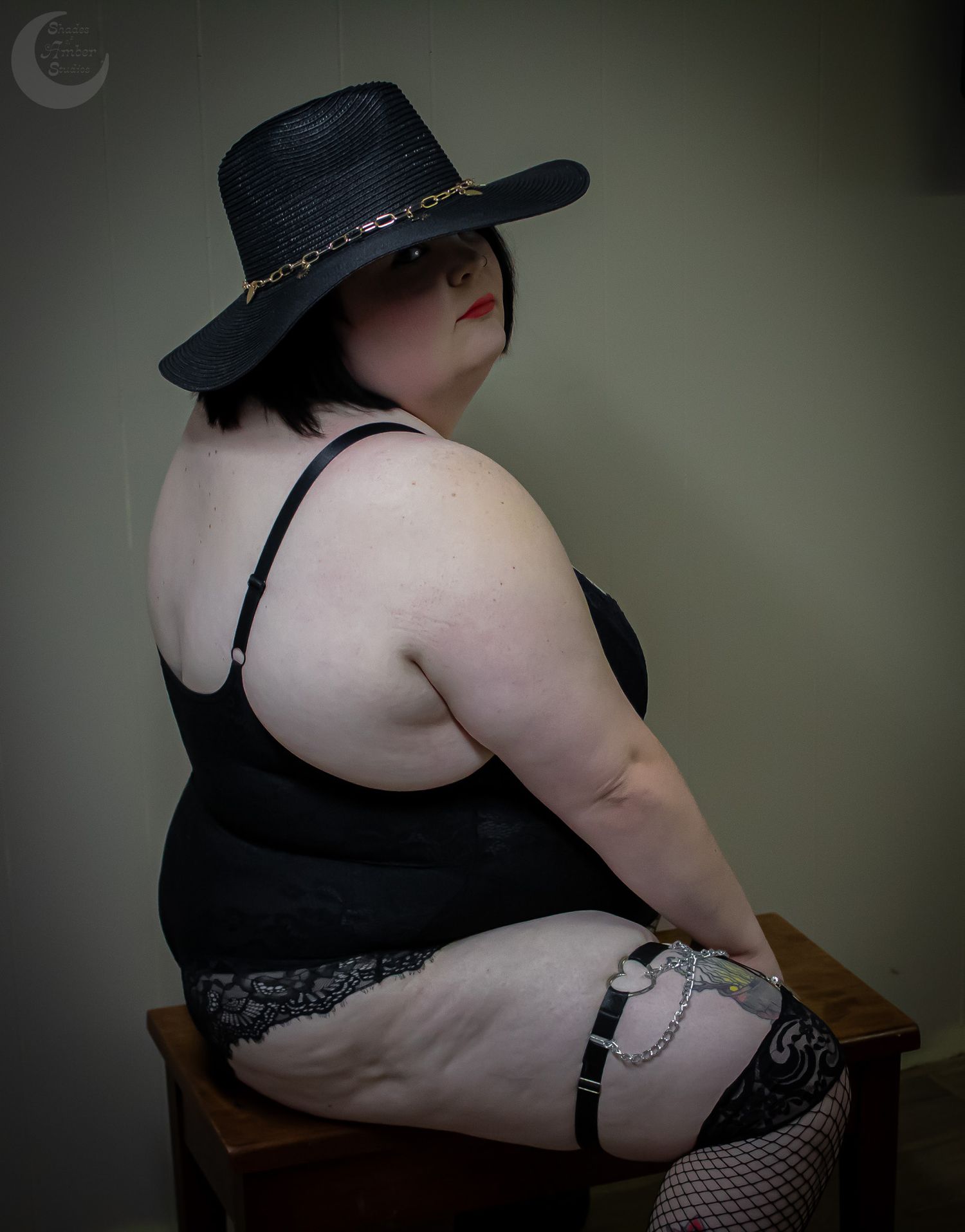 Figure in black hat and lingerie poses on wooden seat in studio setting.