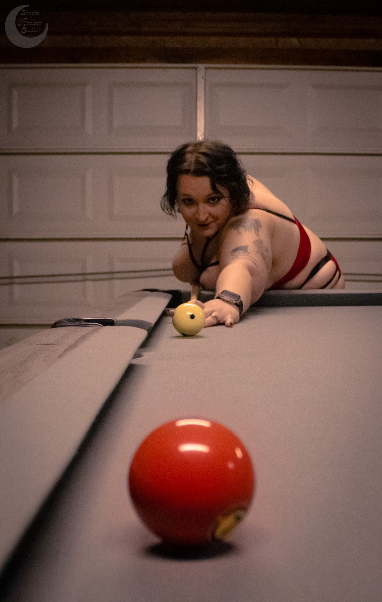 Woman leaning over a pool table aiming at a red billiard ball in a dimly lit garage setting.