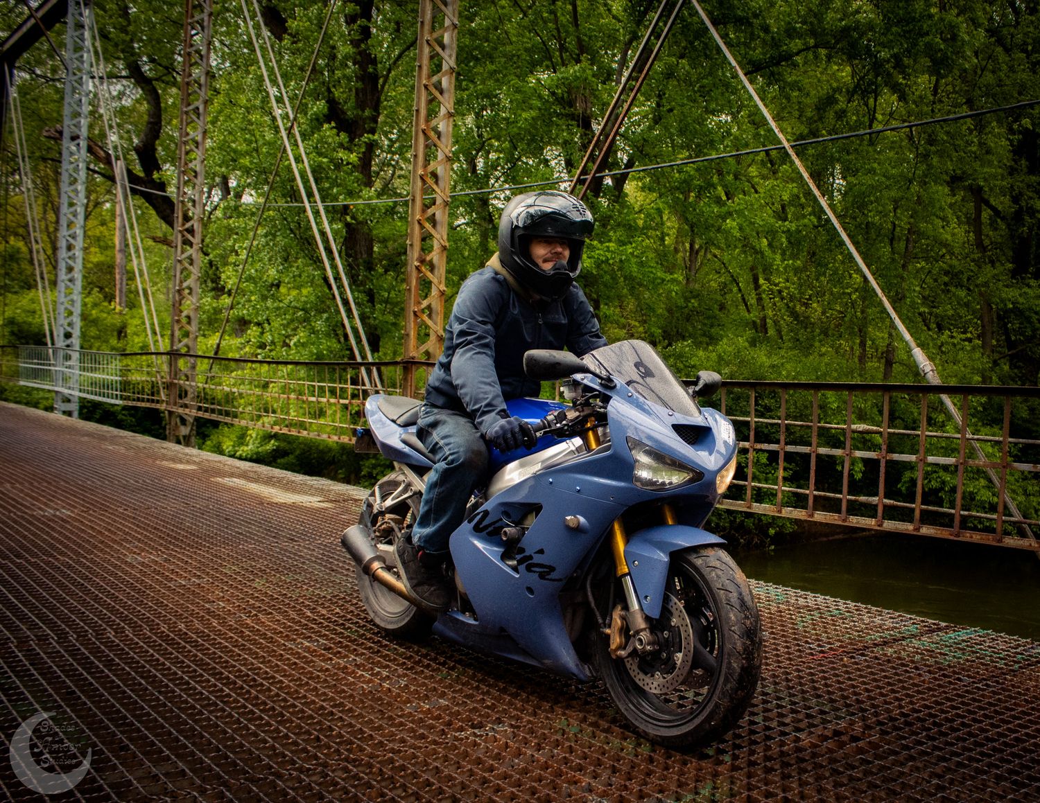 Motorcycle rider crosses vintage wooden bridge on blue sport bike surrounded by green forest scenery.