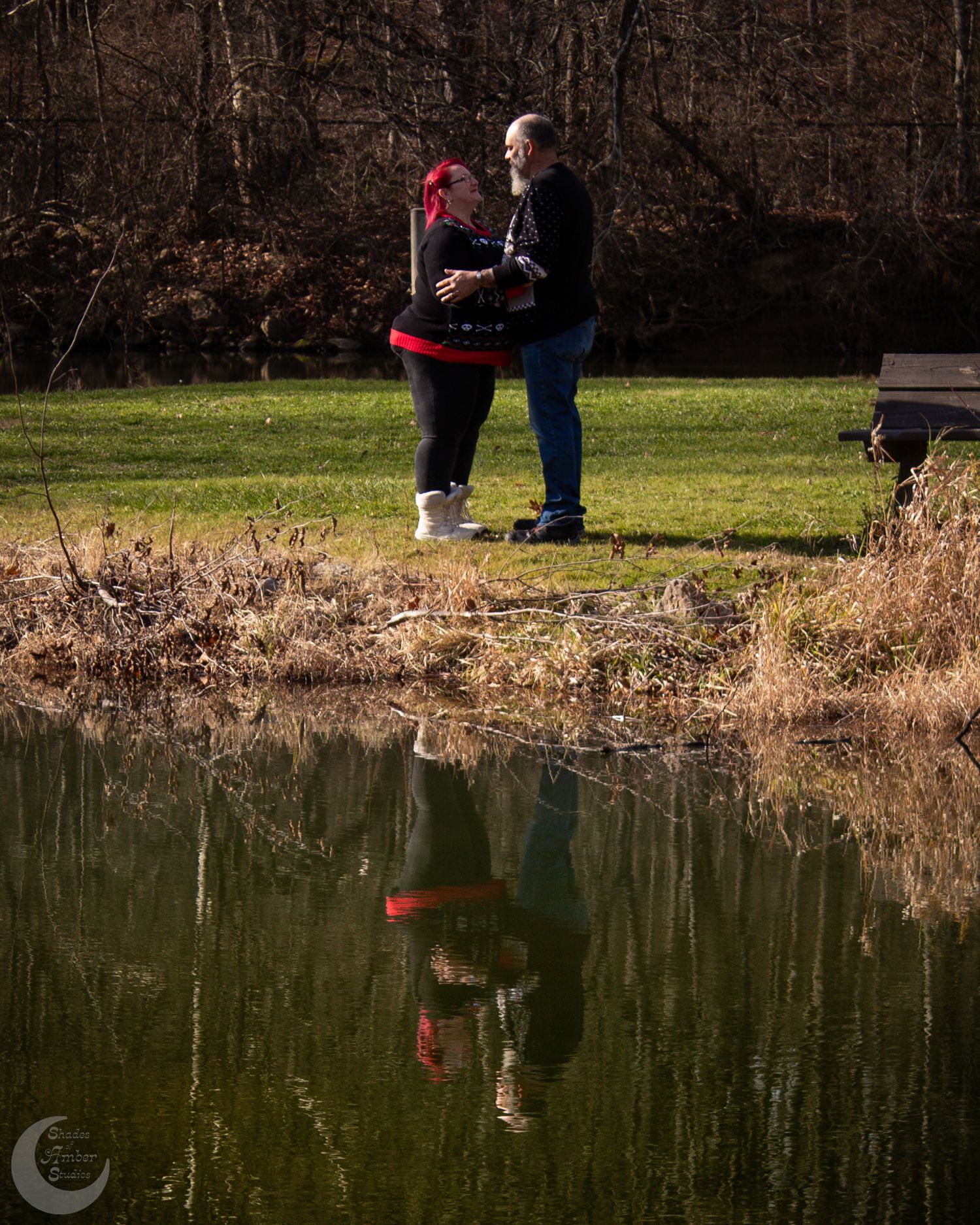 A couple in winter clothing stands by a pond's edge with their reflection mirrored in the still water below.