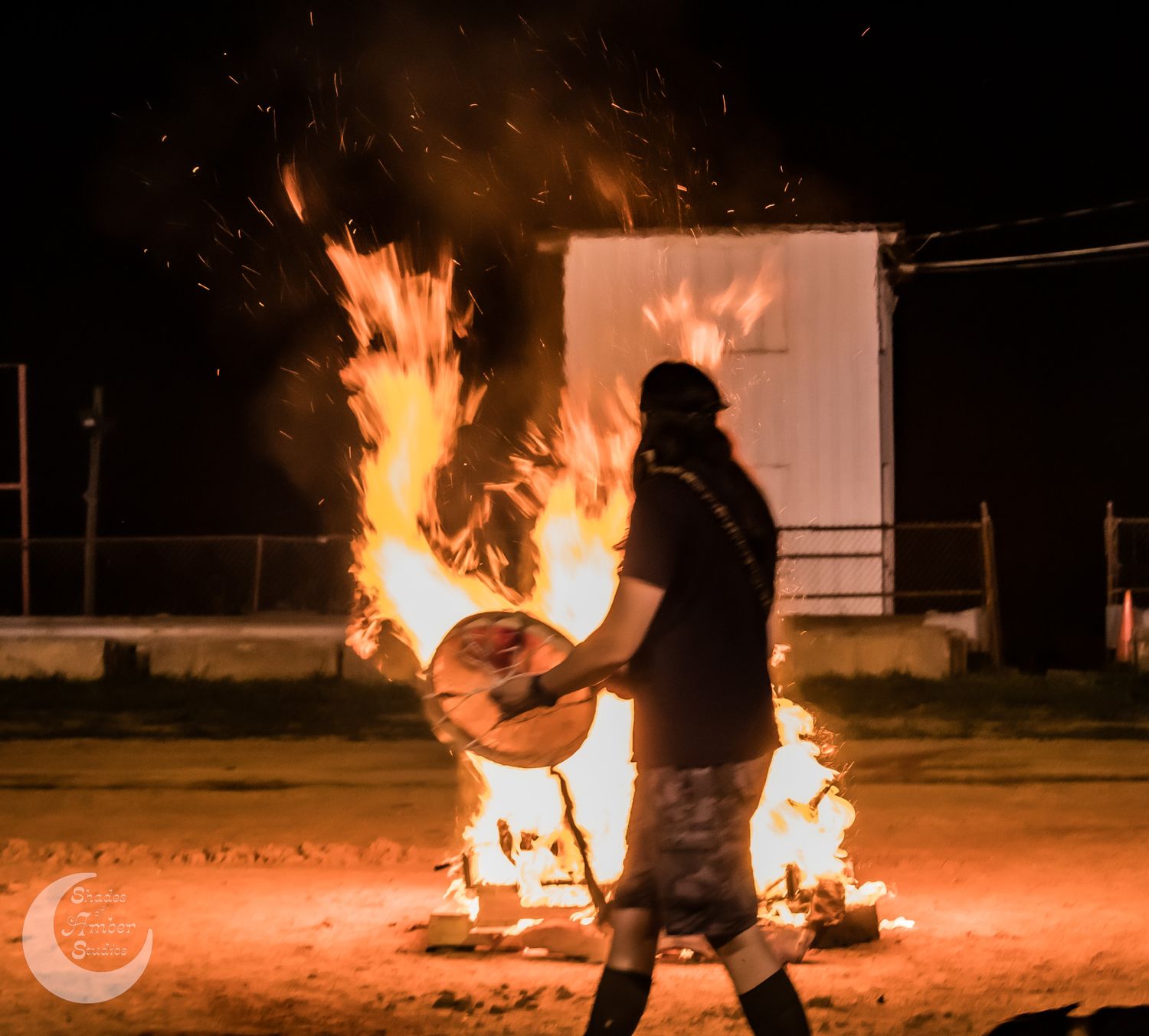 A silhouetted person stands near a bright fire at night with a circular object amidst dramatic orange flames.