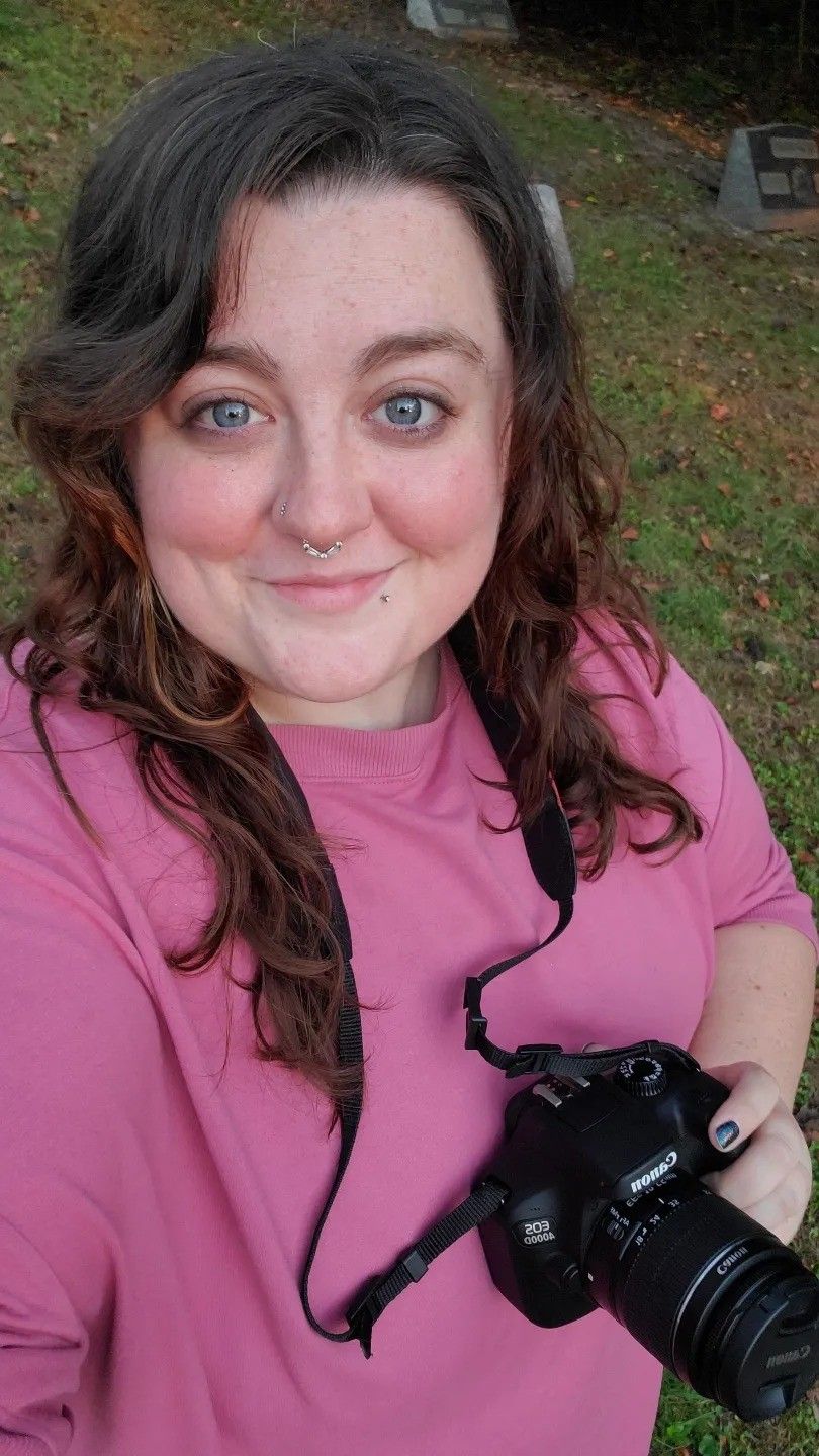 Photographer in pink t-shirt takes selfie with professional camera outdoors.