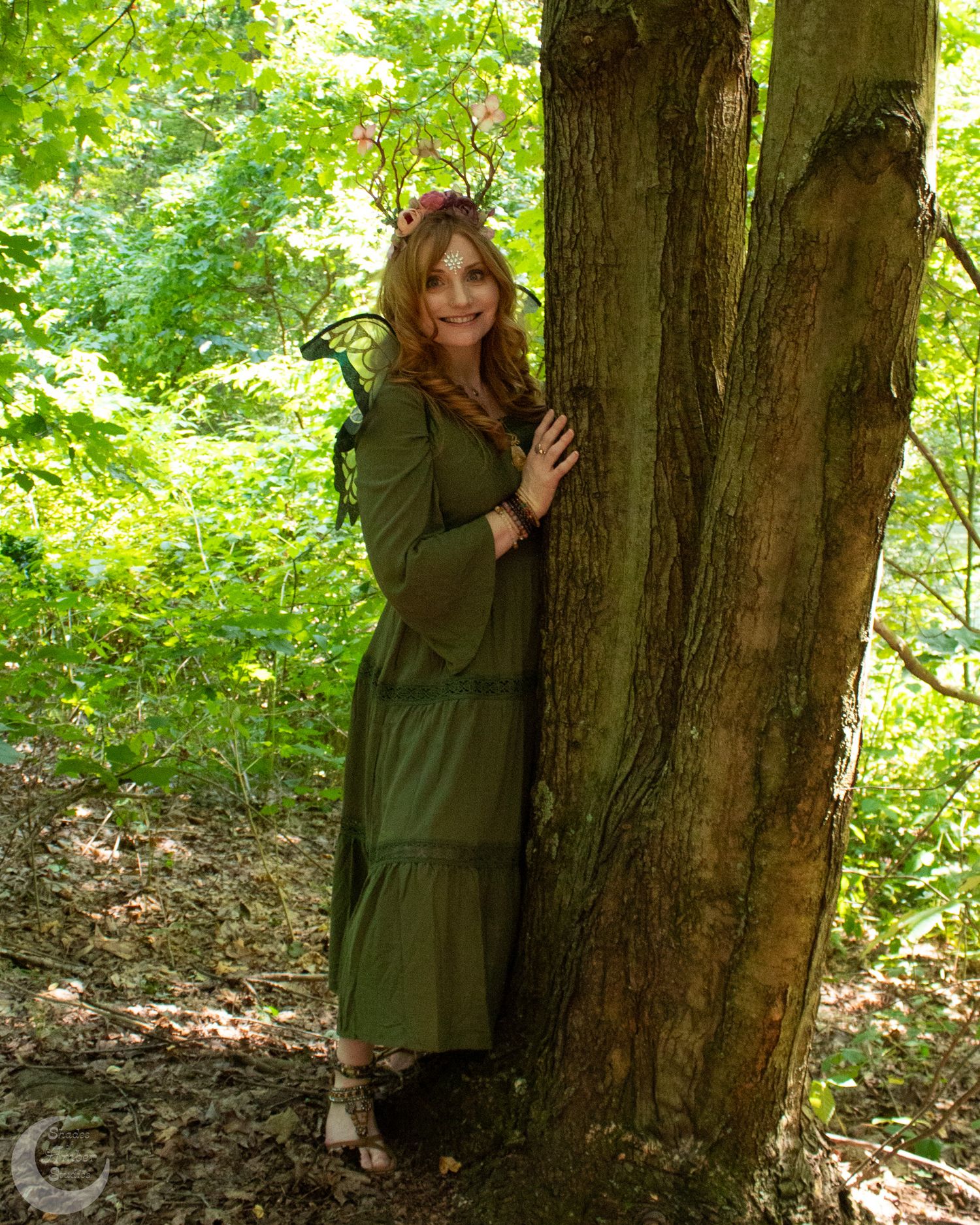 A figure in a green jacket poses playfully behind a large tree trunk in a lush forest setting.