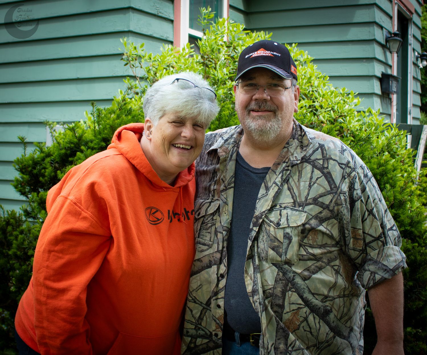 Two people in casual attire stand together outdoors in front of green siding, one wearing camouflage and one in orange.