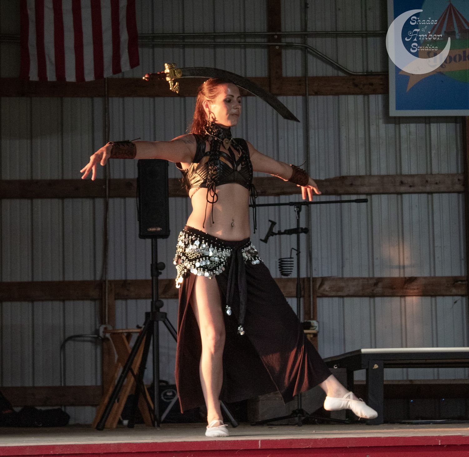 A performer in a belly dance costume dances on stage with dramatic lighting in a rustic indoor venue.