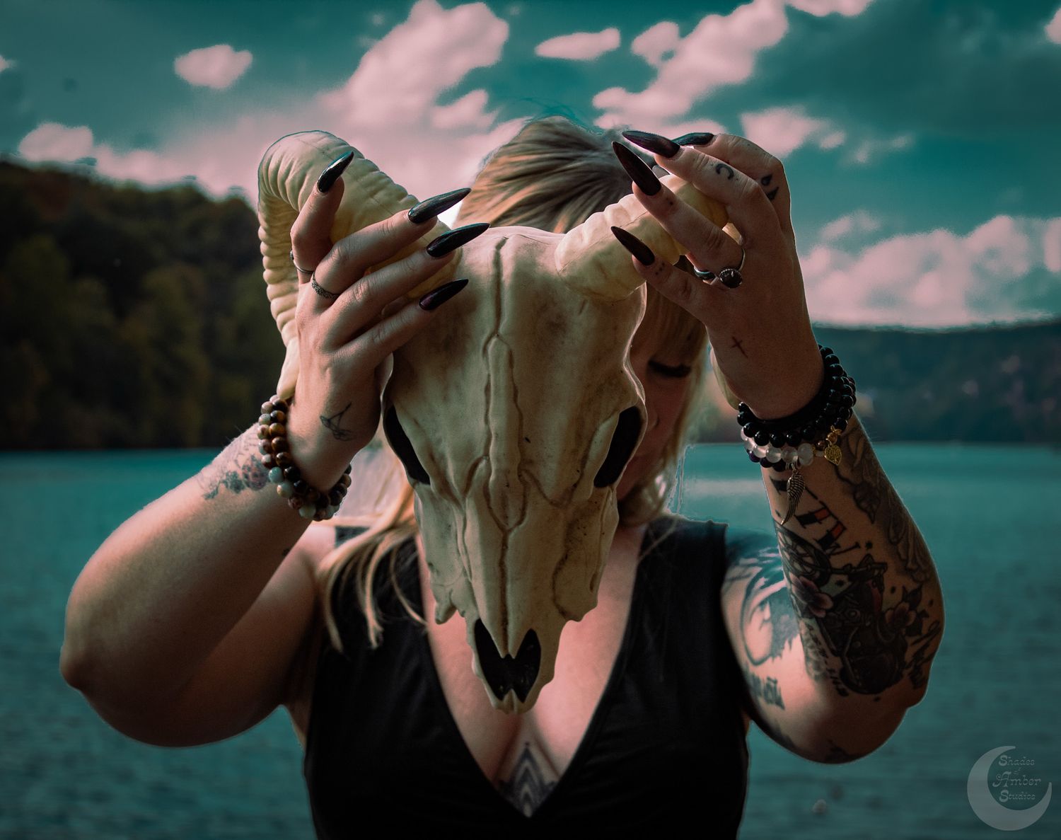 Person holding an animal skull against dramatic cloudy sky near a lake.