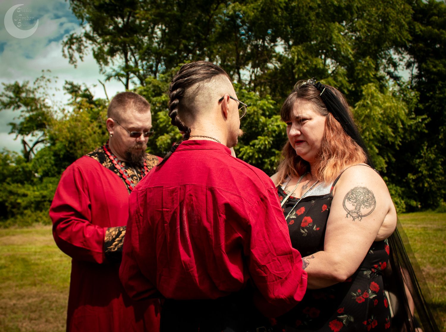 A couple stands outdoors with an officiant in red robes conducting a wedding ceremony in a lush garden setting.