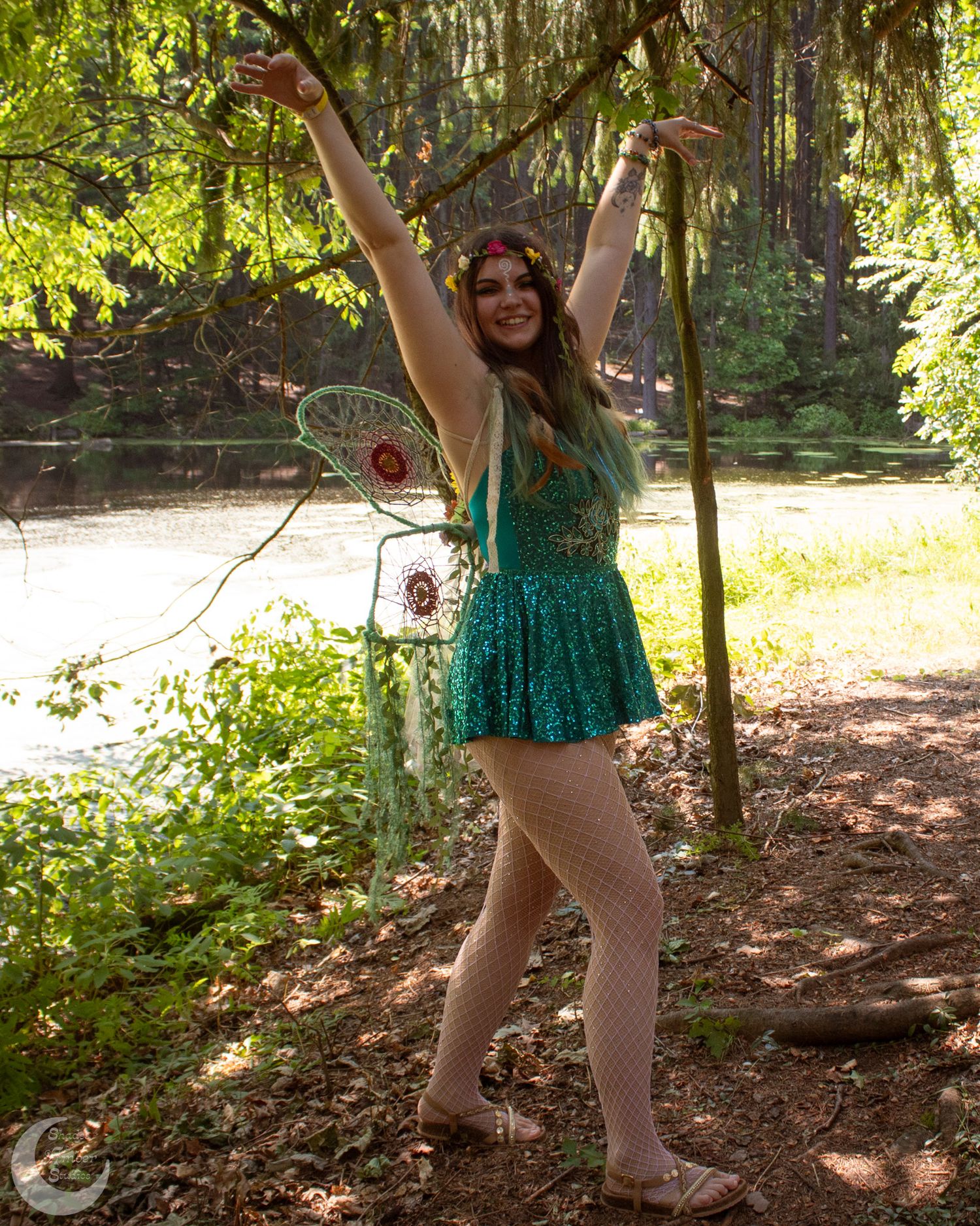 A person wearing a sparkly green fairy costume poses joyfully with raised arms in a sunlit forest setting.