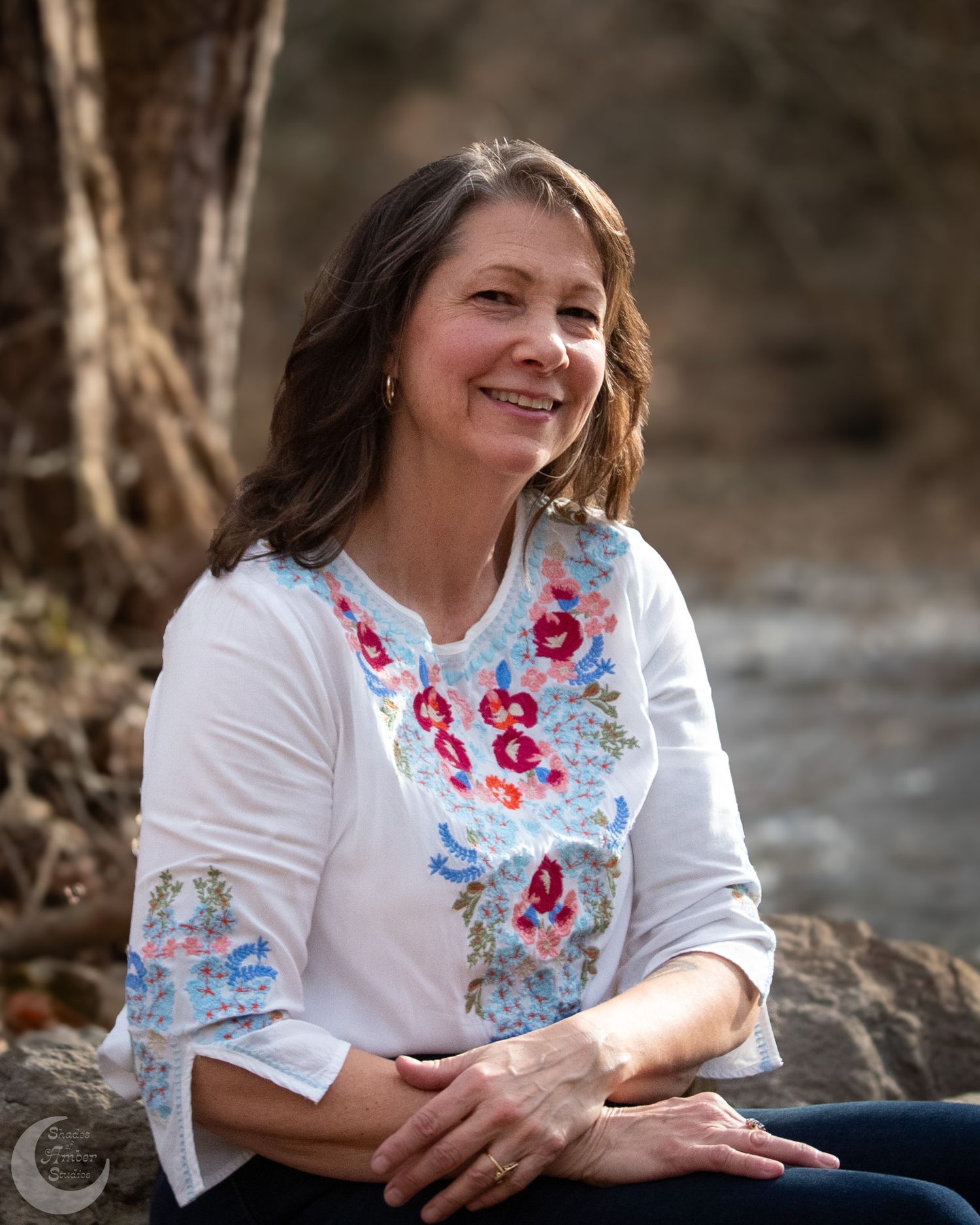 Portrait of someone in embroidered white top sitting by a stream in nature.