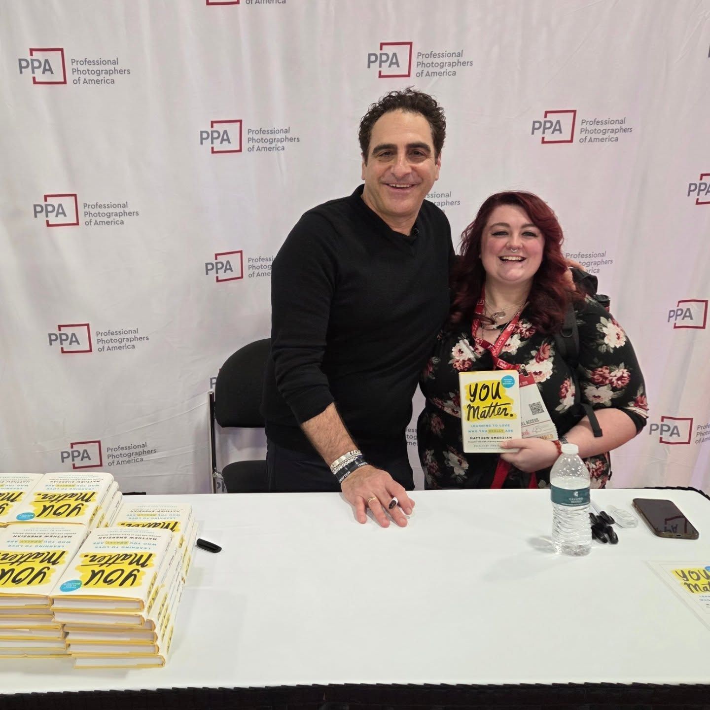 Two people seated at a signing table with promotional materials and books at what appears to be a public event.