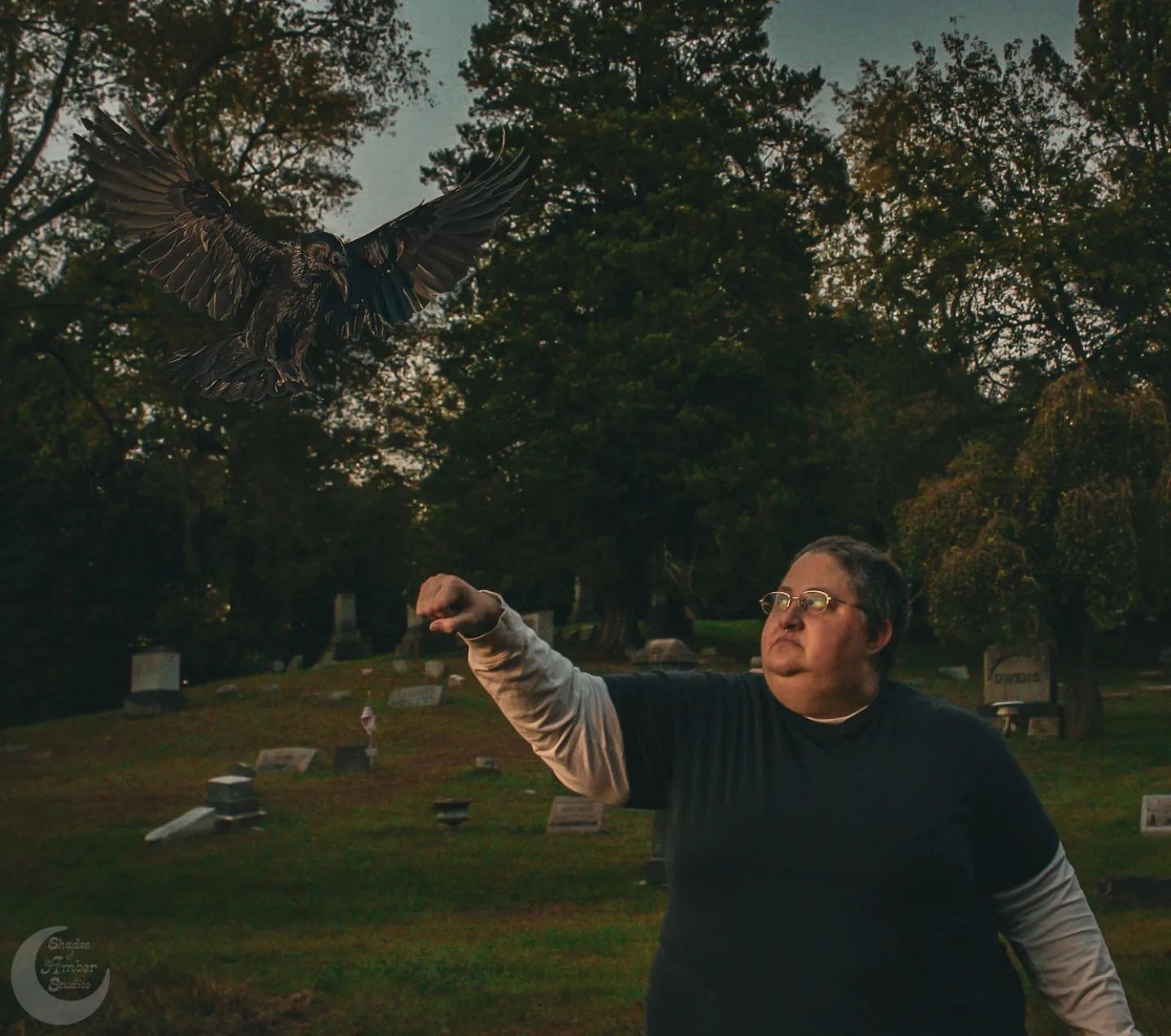 A figure holds up an object in a cemetery at dusk with trees in the background.