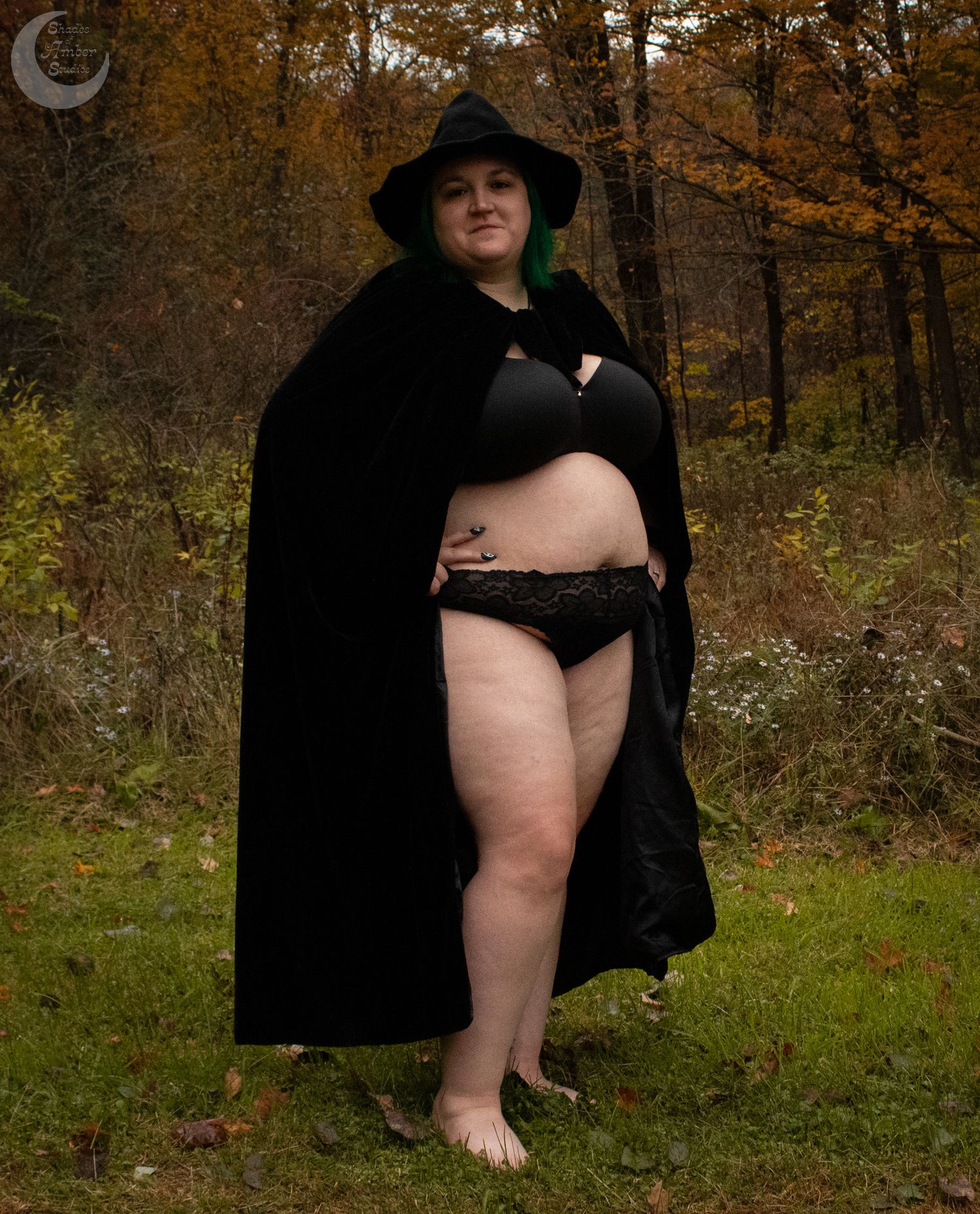 Figure in black cape and lingerie poses barefoot in autumn forest.