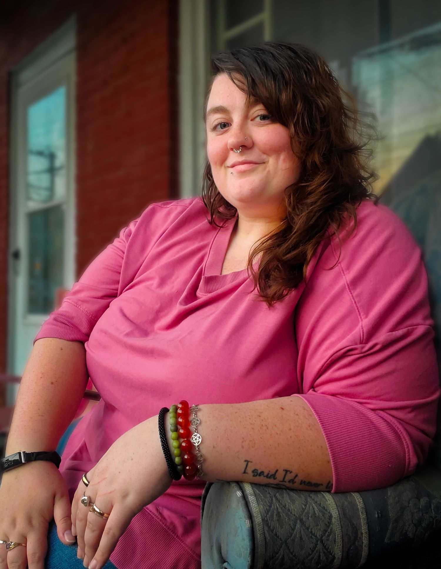 A person wearing a pink blouse with bracelets and rings smiles while sitting outdoors.