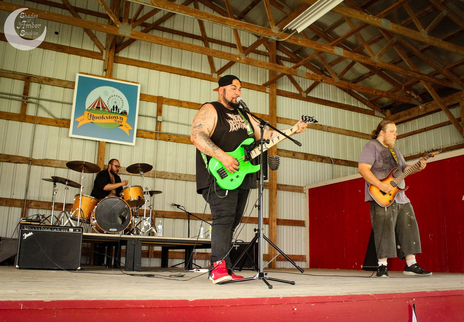 Musicians perform on stage in a wooden barn setting with drums and electric guitars during a live performance.