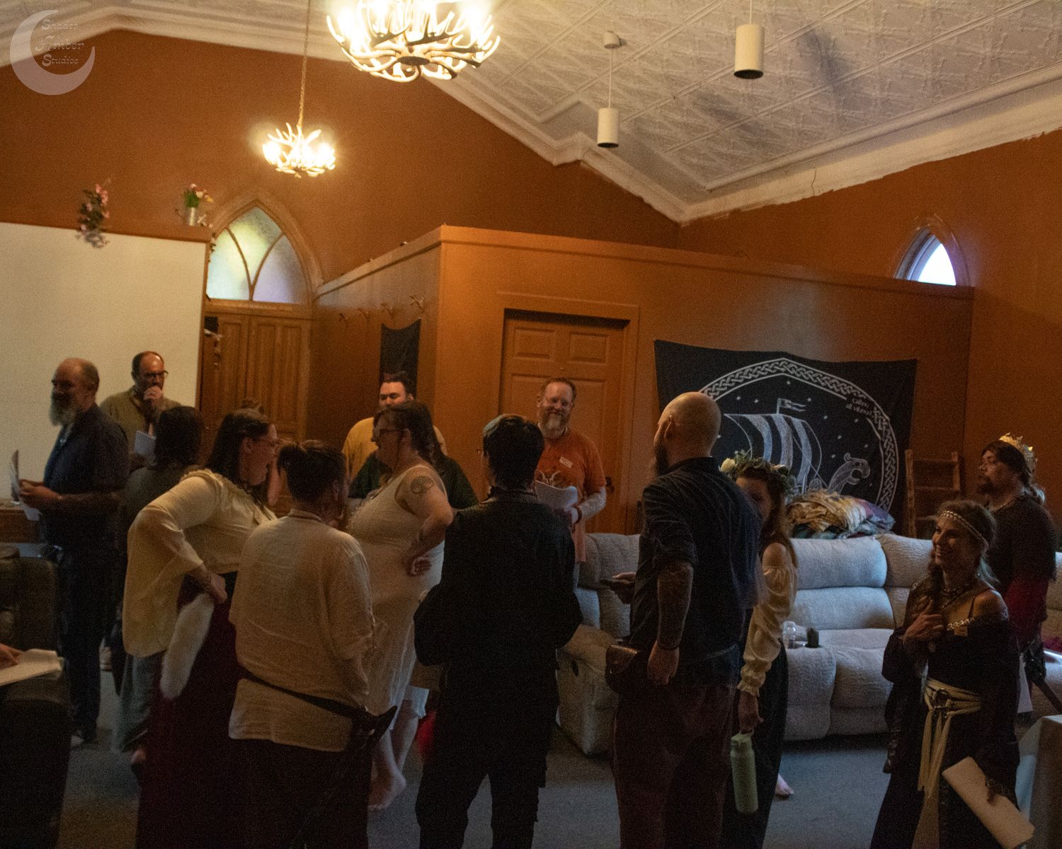 A group of people gather inside a church-like room with arched windows and chandeliers during what appears to be a social event.