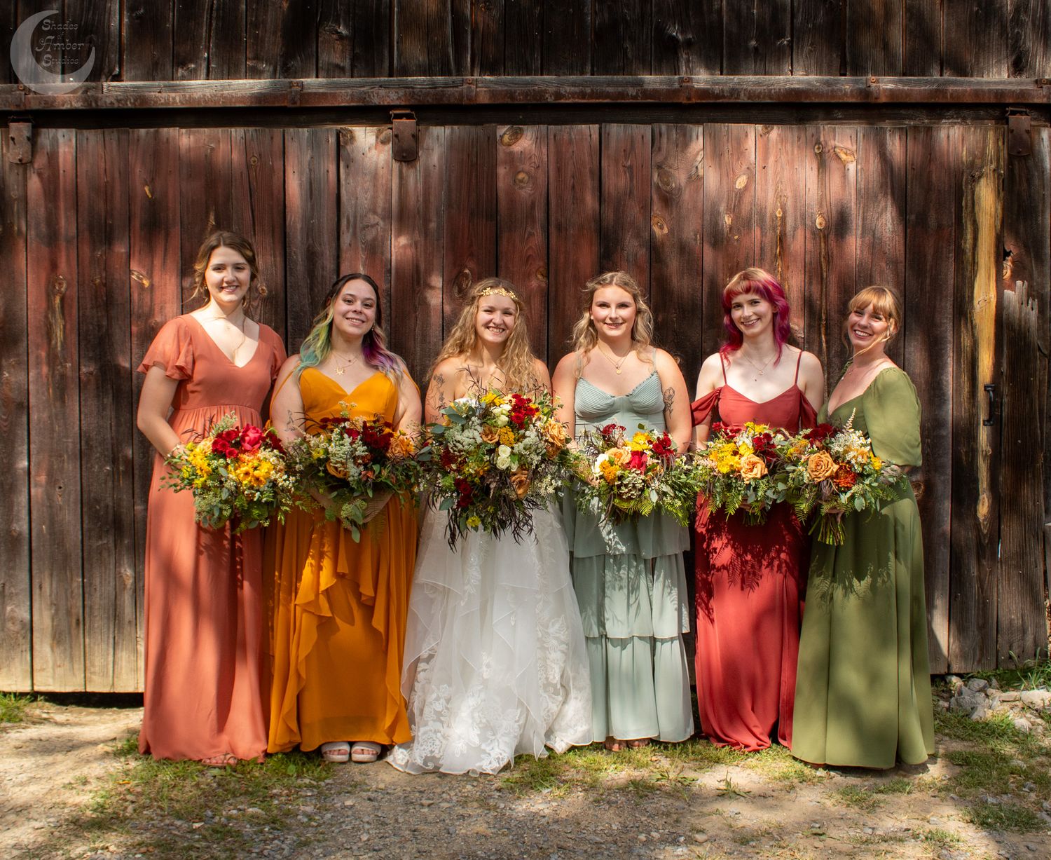 A group in formal wedding attire poses together, with bridesmaids wearing mix-matched dresses in coral and sage tones.