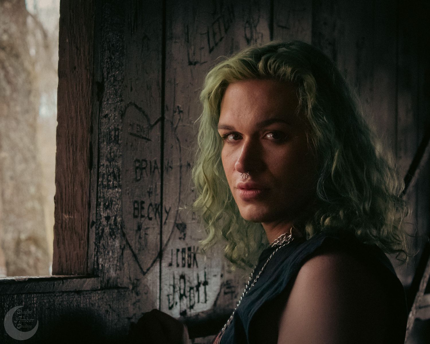 Moody portrait in dark setting with green hair and black tank top against graffitied wall.
