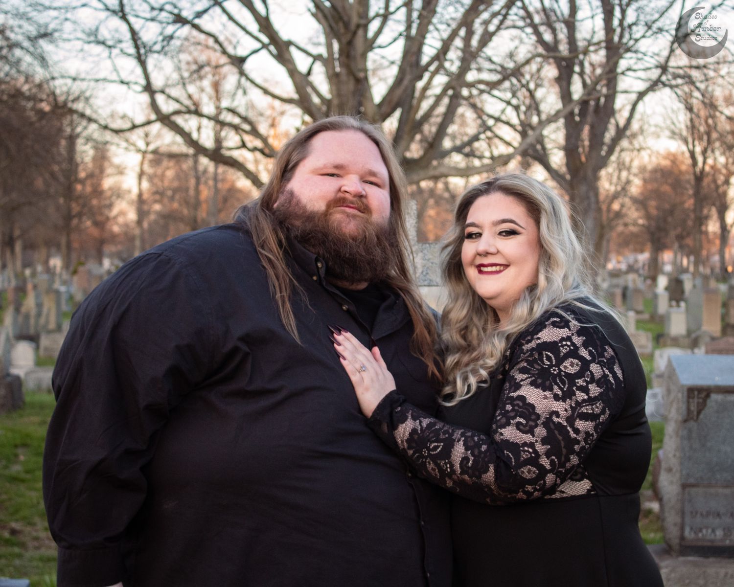 Couple in black outfits posing in a cemetery during golden hour, surrounded by gravestones and bare trees.