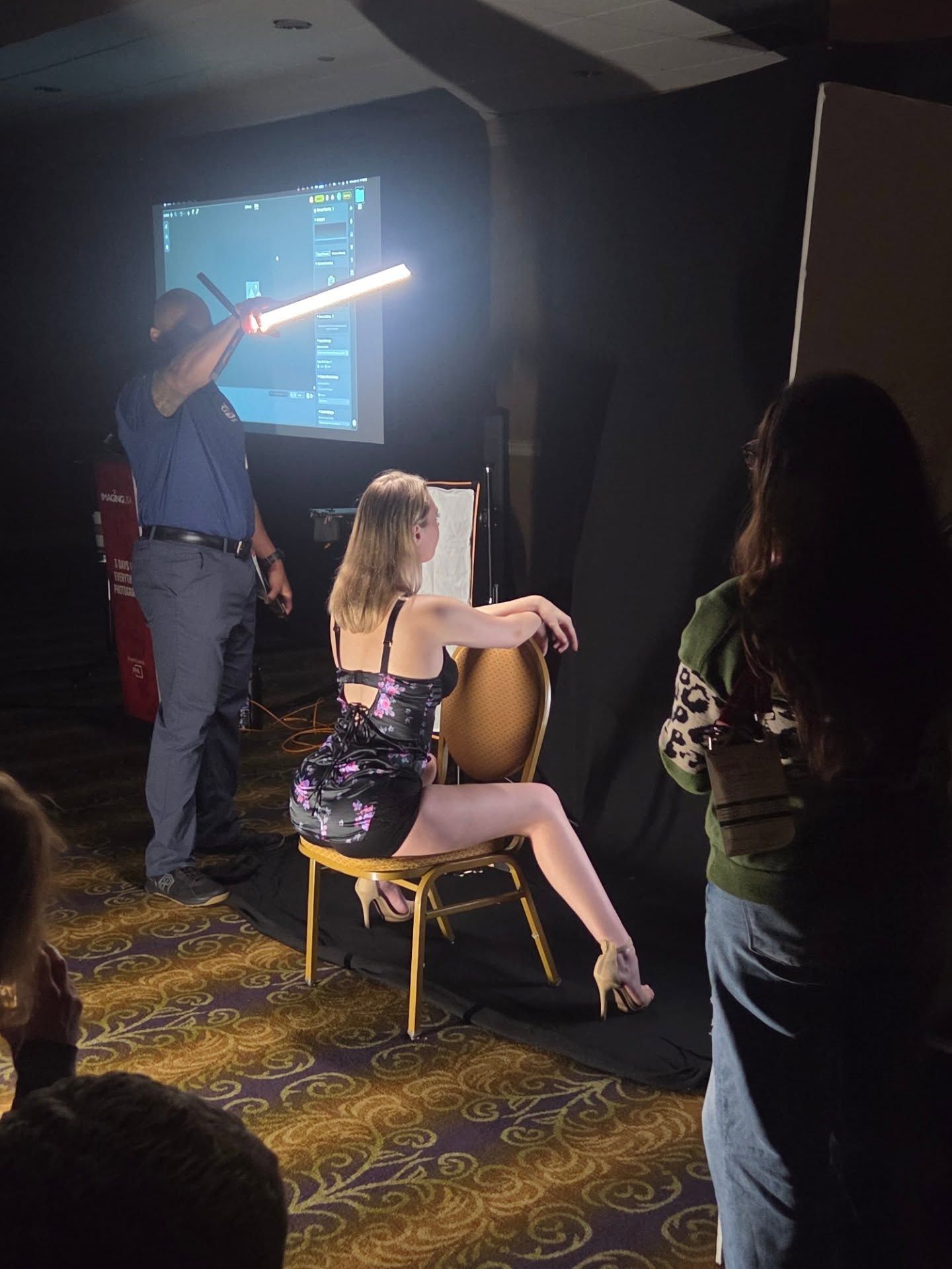 People gather in a dimly lit room with projector screens and gaming equipment visible in the background.