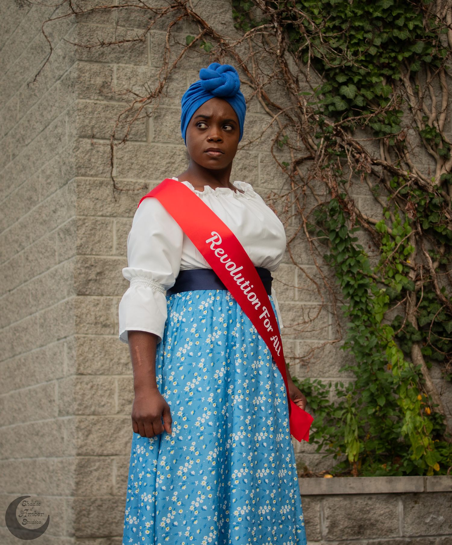 Historical reenactor in blue and white period dress with red sash poses against stone wall with climbing ivy.