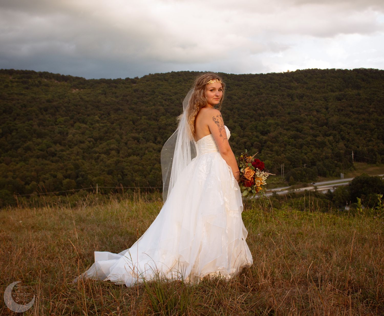 A bride in a flowing white gown walks through a beautiful autumn meadow with mountains in the background at sunset.