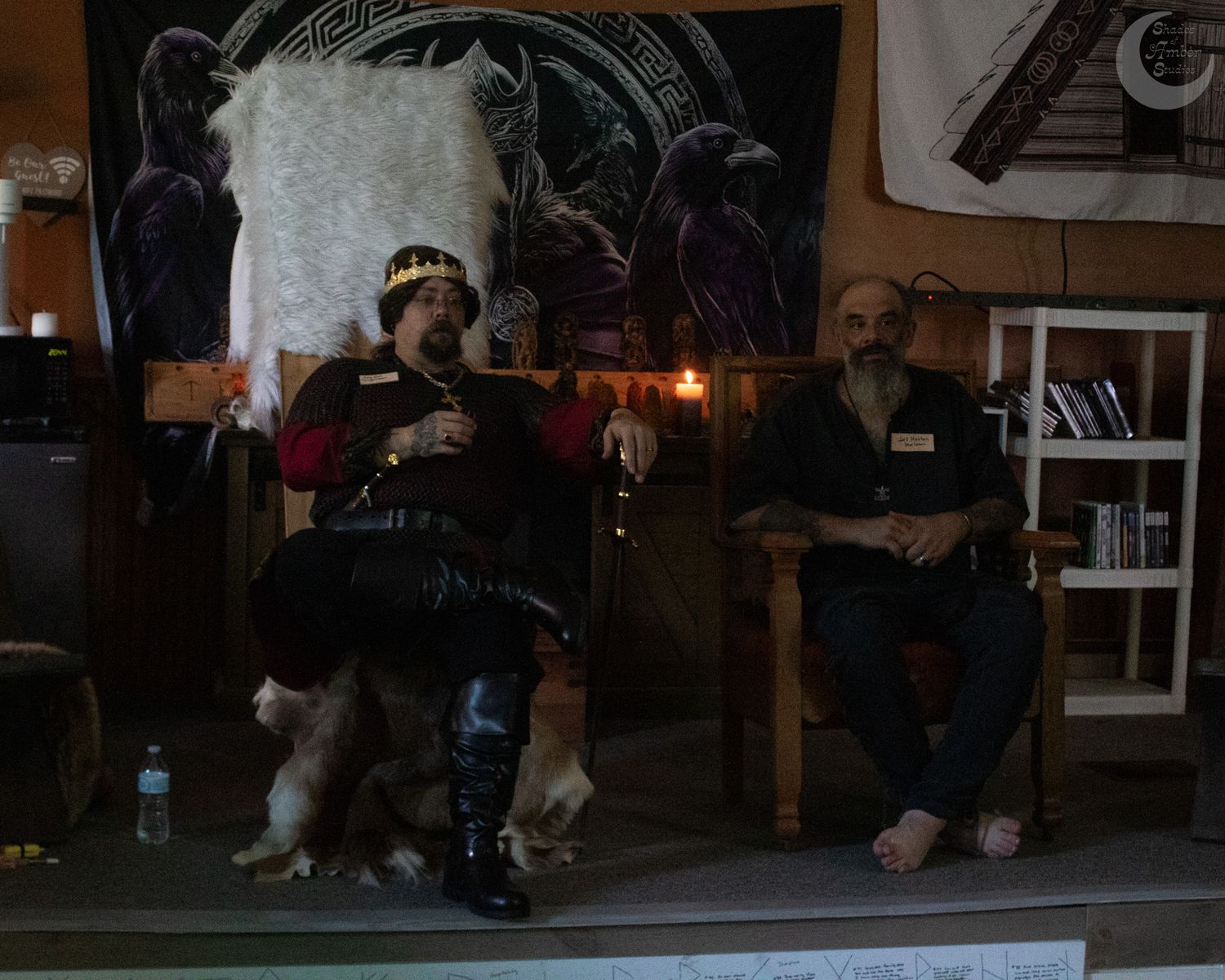 Two people sit in a dark room with candles lit, surrounded by artistic wall decorations and dim ambient lighting.
