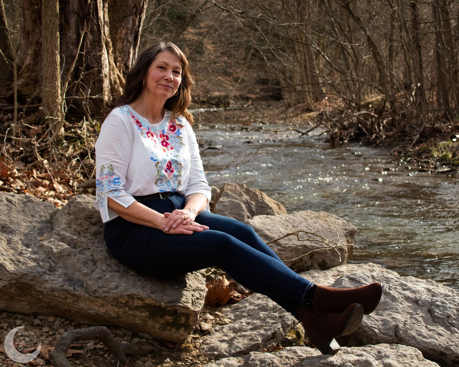 Casual portrait by a stream showing someone relaxing on rocks in embroidered top and jeans.