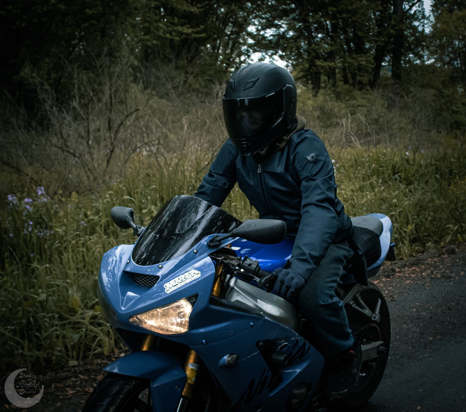 A motorcyclist in dark protective gear rides a blue sportbike on a rural road surrounded by foliage.
