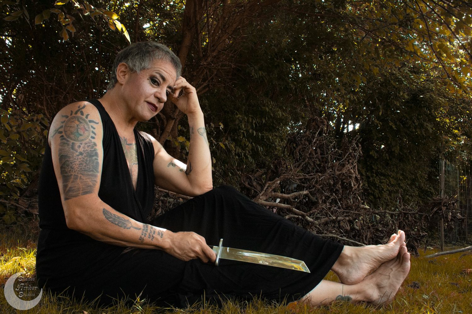 Artistic portrait of tattooed person in black clothing resting on grass in natural lighting.