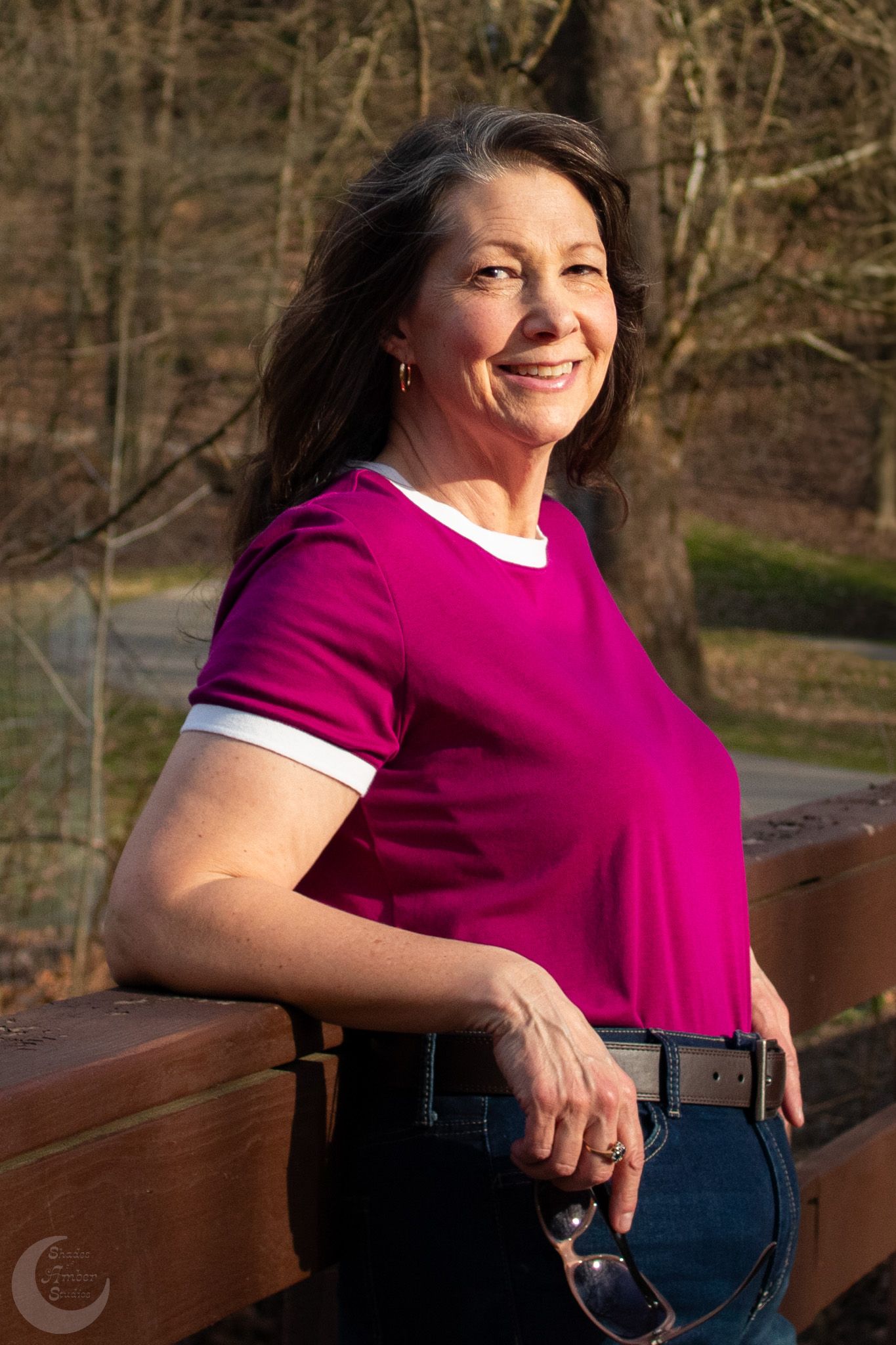 Side-lit portrait showing confident pose in magenta shirt against wooded background.