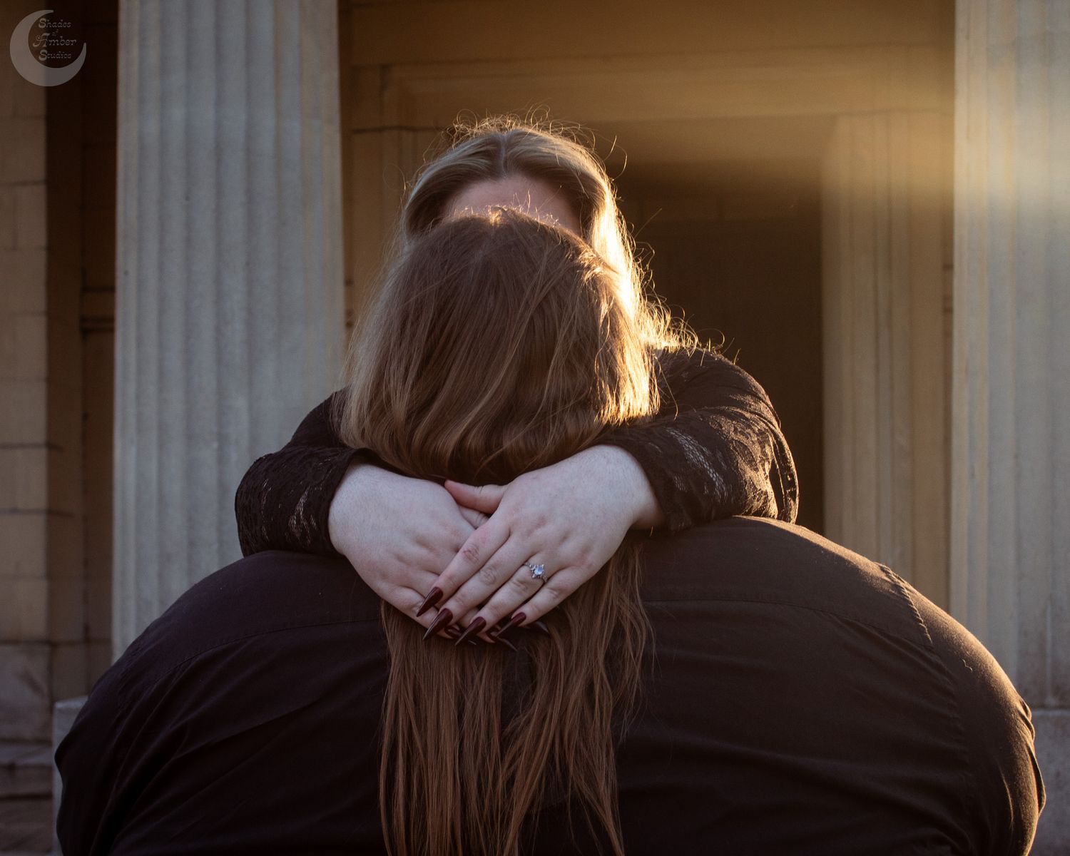 Couple embracing warmly in golden sunset light near stone columns, woman with long hair hugging man from behind.