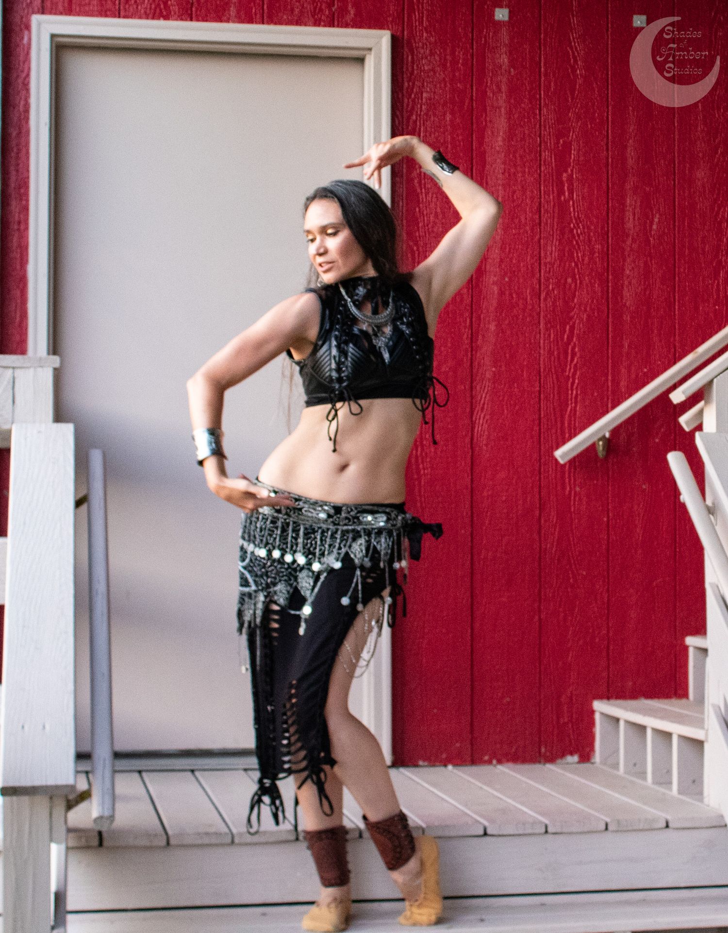 A dancer poses in a black tribal belly dance costume with coin belt and fringe against a red and white backdrop.