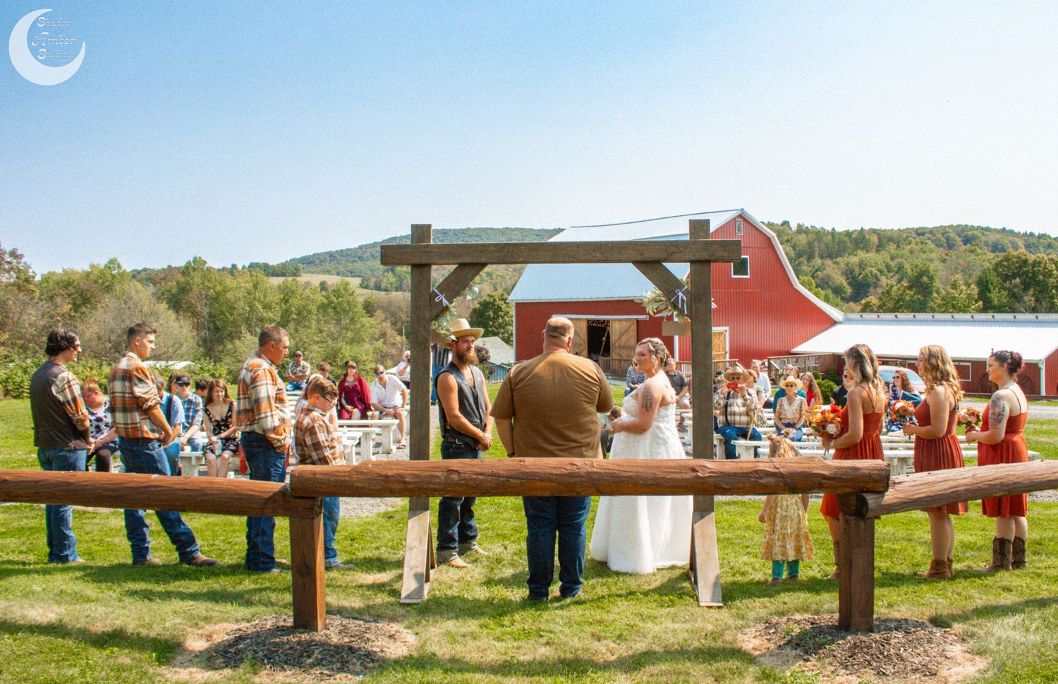 Wedding ceremony guests gathered at rustic outdoor venue with wooden benches and red barn in background.