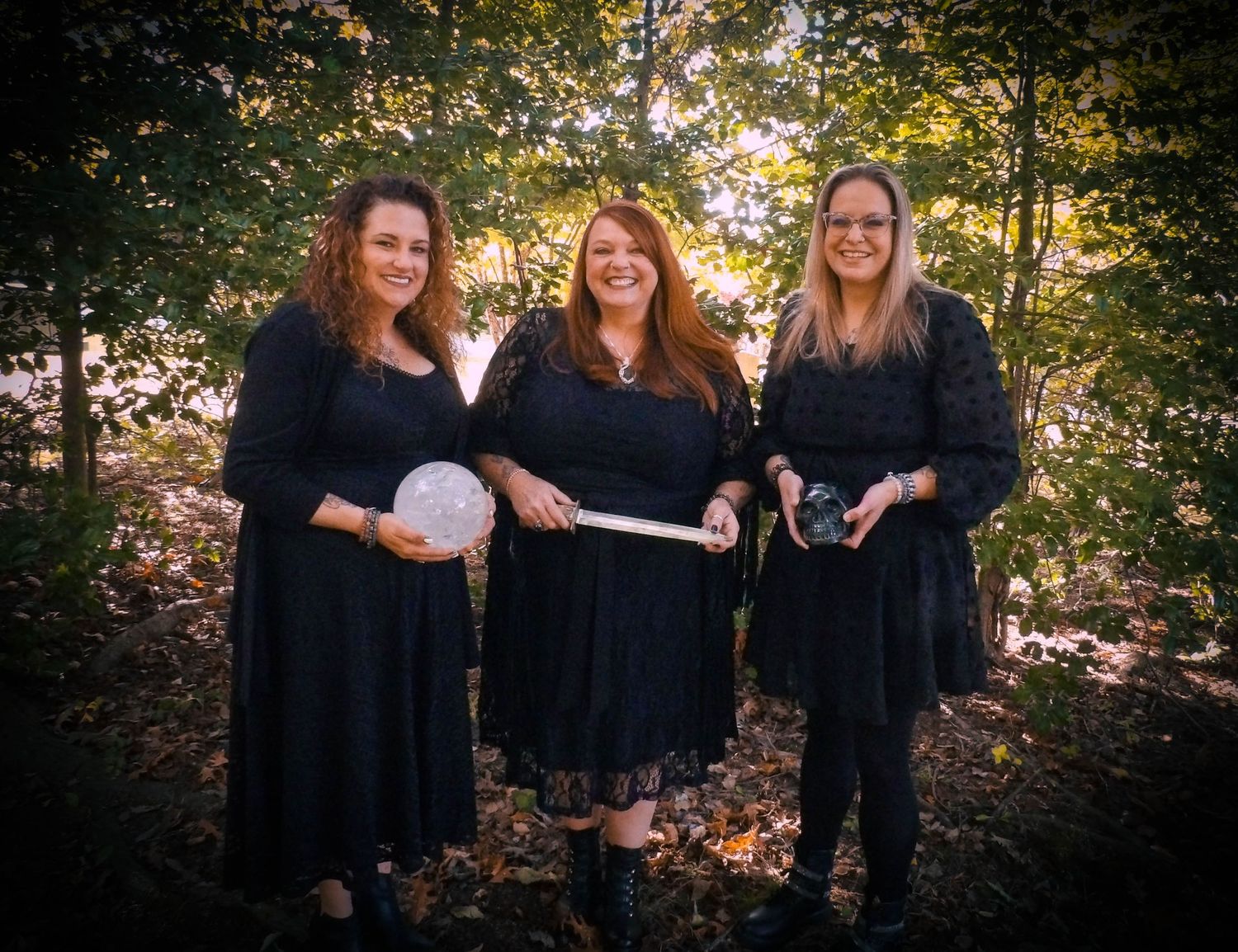 Three people in black dresses holding props in autumn woodland setting.