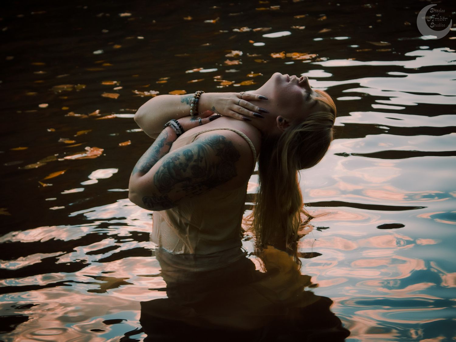 Artistic photo of person floating in dark water with tattoos and reflective ripples at sunset.