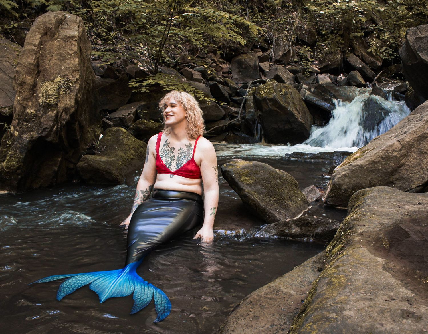 A mermaid in a blue tail poses on rocky waterfall outcropping.