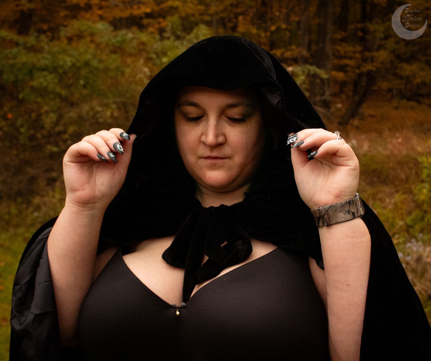 Person adjusting black hooded cape in moody forest portrait.