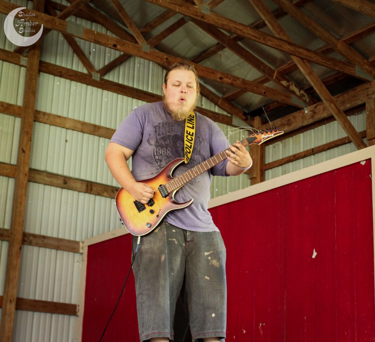Musician performs passionately on electric guitar in rustic barn setting with red backdrop.
