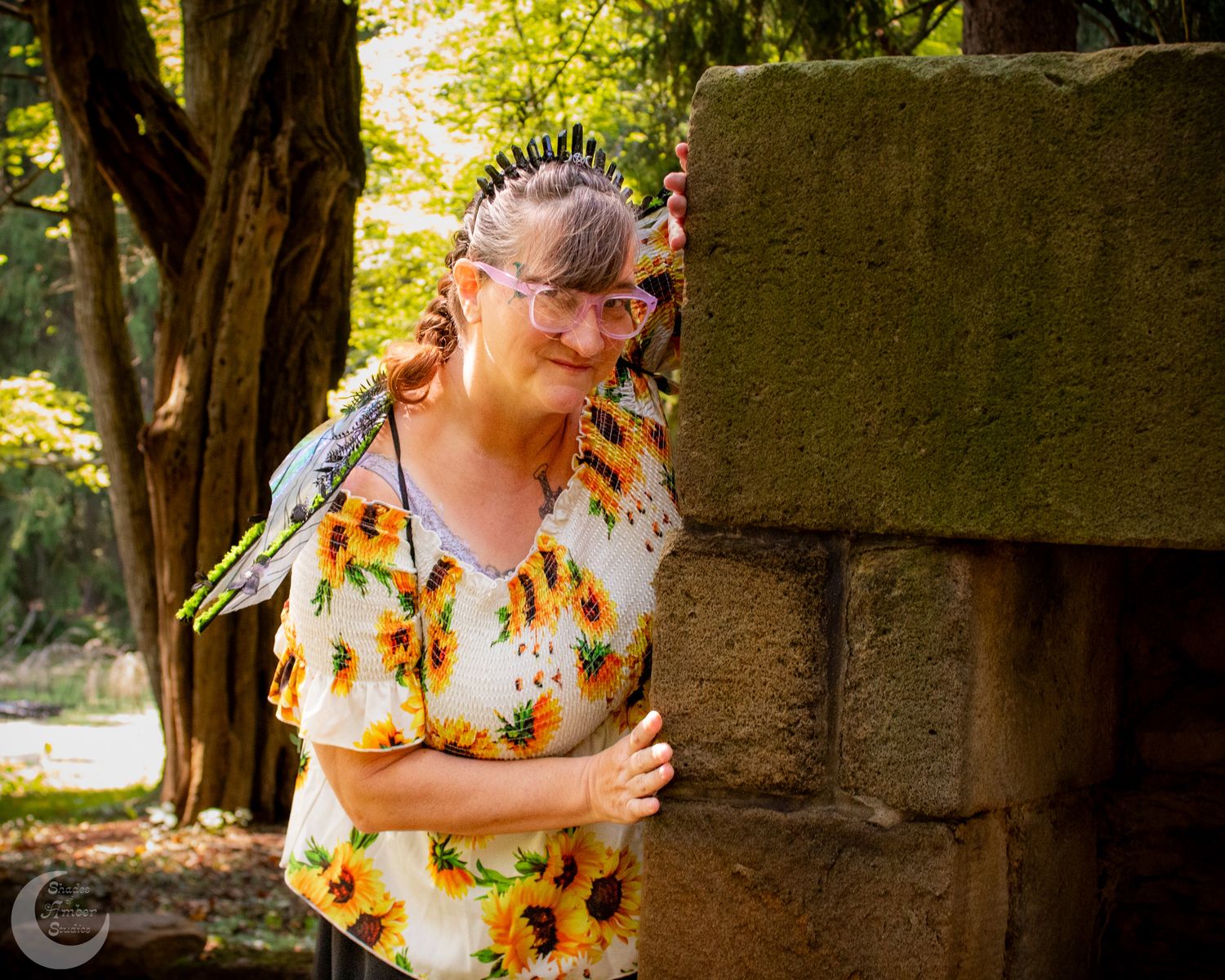 Woman in yellow floral dress poses against a stone wall in a garden setting.