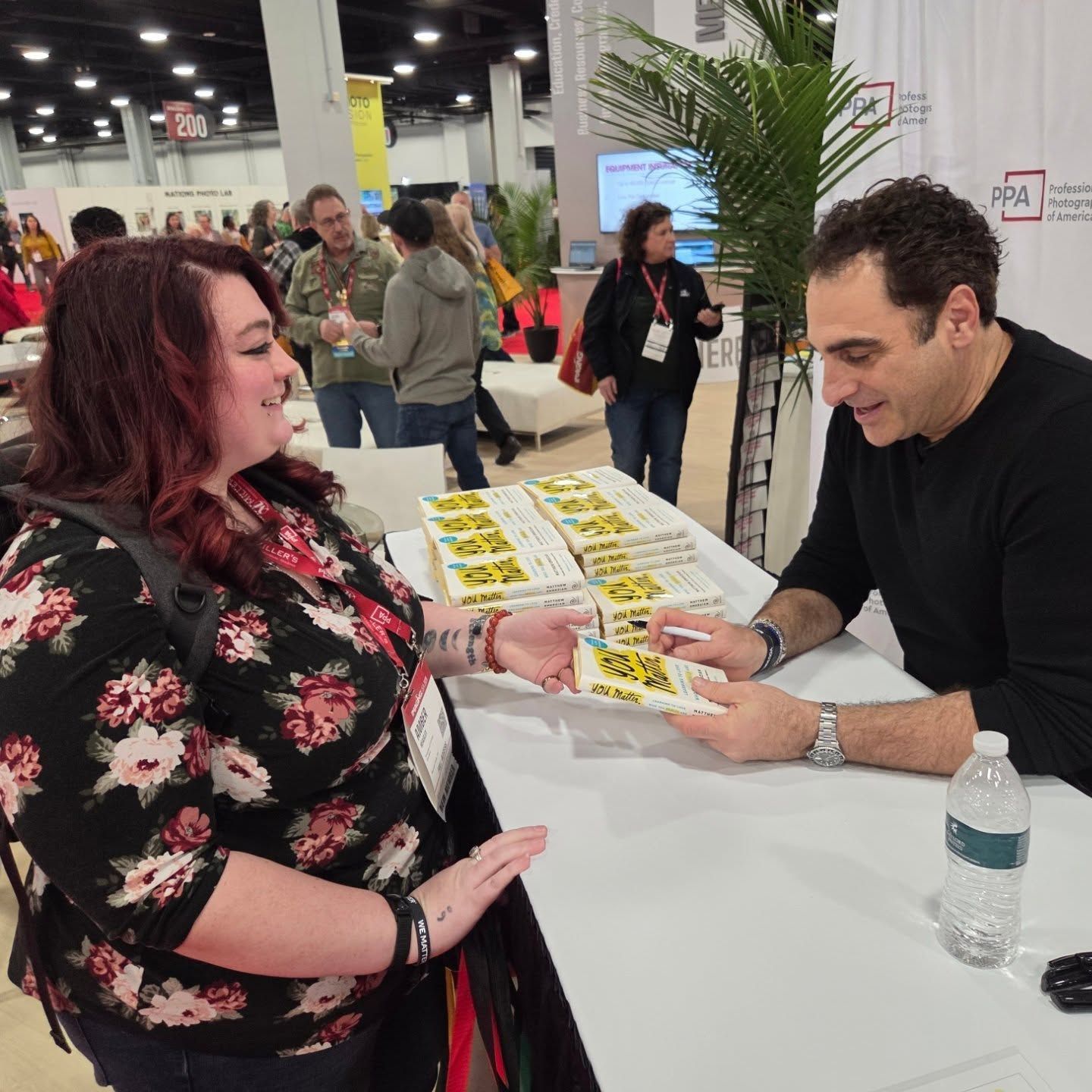 A person in a floral dress signs books at an event while attendees wait in line at a table.