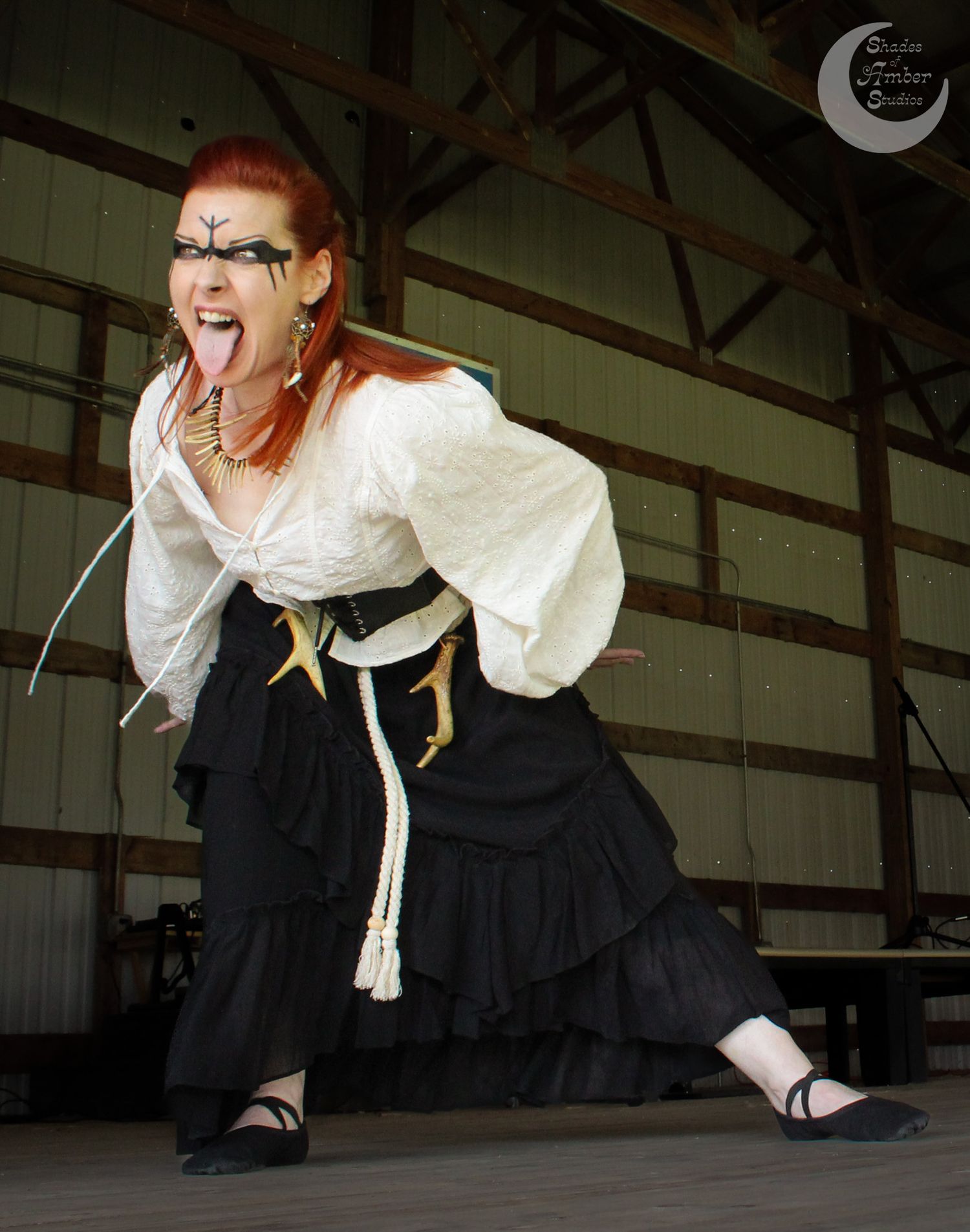 Costumed performer in a white blouse and black skirt strikes an expressive pose with tongue out on stage indoors.