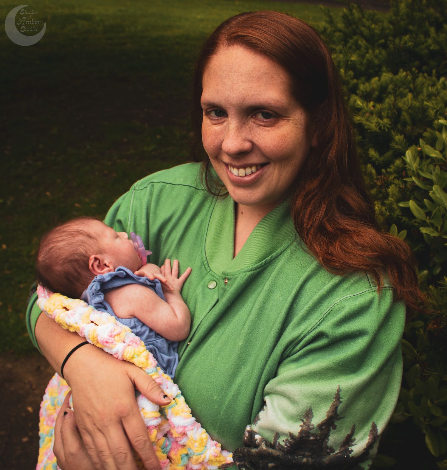 A person in a green shirt holds and comforts a small baby wearing a floral outfit in an outdoor garden setting.